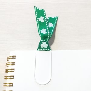 May include: A white metal bookmark with a green ribbon with white shamrock designs. The ribbon is tied in a bow at the top of the bookmark.