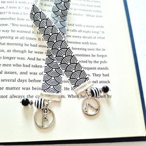 May include: Two fabric bookmarks featuring a black and white repeating wave pattern. Each bookmark is finished with a silver-toned metal charm depicting a person in a yoga pose, accented by black and white striped and solid black spherical beads.
