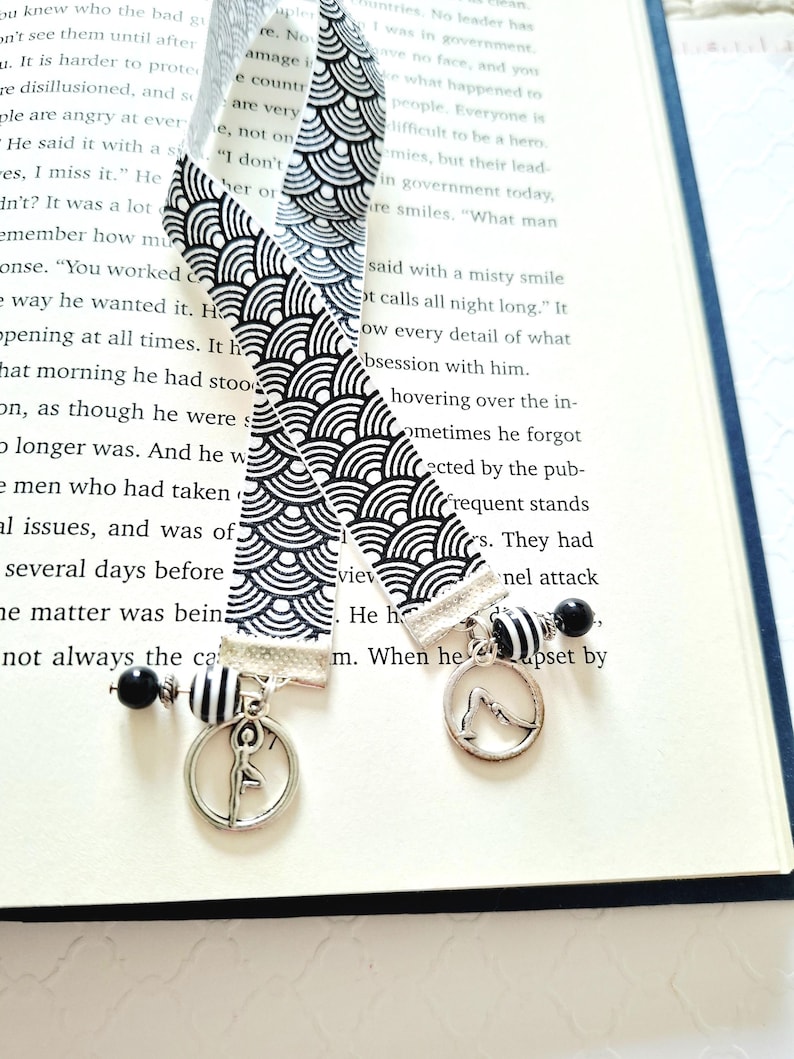 May include: Two fabric bookmarks featuring a black and white repeating wave pattern. Each bookmark is finished with a silver-toned metal charm depicting a person in a yoga pose, accented by black and white striped and solid black spherical beads.
