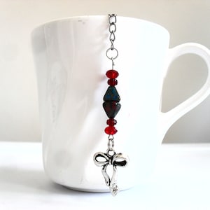 May include: A silver tea infuser with a red and blue beaded chain and a silver bow charm. The infuser is hanging from the rim of a white teacup.