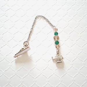 May include: A silver-toned metal clip attached to a short chain featuring alternating green and light brown faceted beads. The chain ends with a small, detailed silver-toned teapot charm. The design is functional and decorative for garment use.
