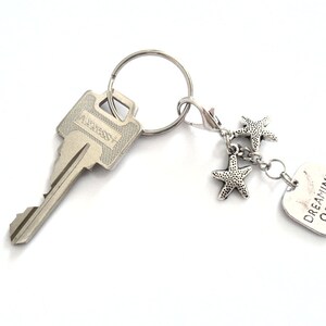 May include: Silver-toned keychain with a key, a lobster clasp, and a charm. The charm features three starfish and a rectangular tag with the words "DREAMING OF THE SEA".