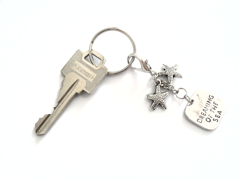 May include: Silver-toned keychain with a key, a lobster clasp, and a charm. The charm features three starfish and a rectangular tag with the words "DREAMING OF THE SEA".