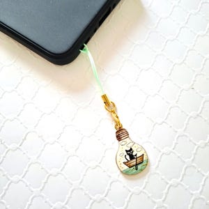 May include: A black smartphone is connected to a light green lanyard with a gold-colored clasp. The lanyard is attached to a charm in the shape of a lightbulb. Inside the lightbulb is a black cat in a boat.