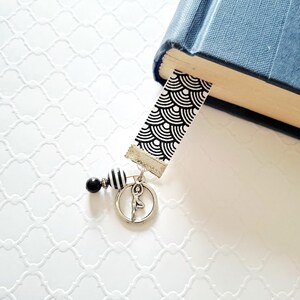 May include: A bookmark featuring a black and white geometric fan pattern on a fabric strip. Attached to the bottom is a silver-toned metal charm of a person in a yoga pose, along with a small black bead and a black and white striped bead.