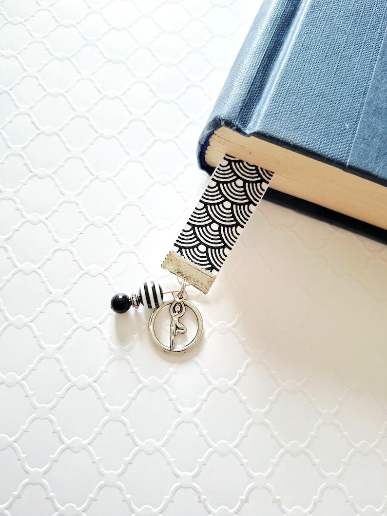 May include: A bookmark featuring a black and white geometric fan pattern on a fabric strip. Attached to the bottom is a silver-toned metal charm of a person in a yoga pose, along with a small black bead and a black and white striped bead.