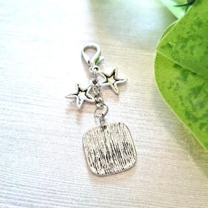 May include: Silver-toned charm with a lobster clasp, two star charms, and a textured square pendant. The charm is designed for jewelry making or as a decorative accessory. The background is a light wood grain.