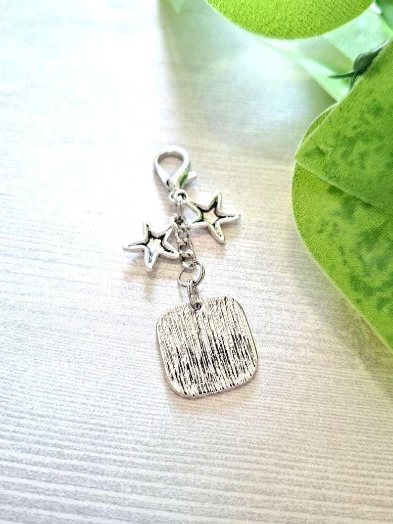 May include: Silver-toned charm with a lobster clasp, two star charms, and a textured square pendant. The charm is designed for jewelry making or as a decorative accessory. The background is a light wood grain.