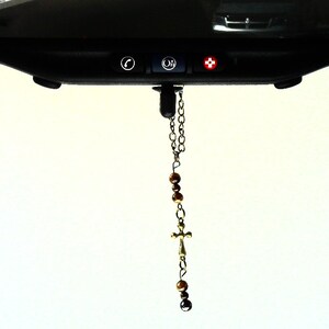 May include: A hanging car accessory featuring a silver-toned metal chain, brown tiger eye beads, and a small metal cross. The ornament is suspended from a black vehicle rearview mirror that includes three buttons with phone, OnStar, and medical icons.