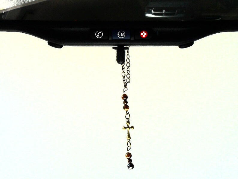 May include: A hanging car accessory featuring a silver-toned metal chain, brown tiger eye beads, and a small metal cross. The ornament is suspended from a black vehicle rearview mirror that includes three buttons with phone, OnStar, and medical icons.