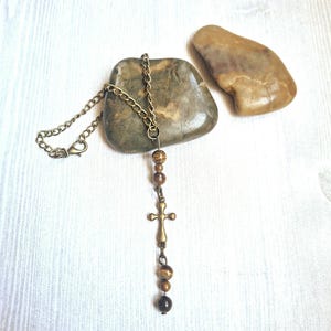 May include: A necklace featuring a bronze-toned metal chain and a small cross pendant. The design includes several round, brown, and tan tiger eye beads arranged above and below the cross, finished with a lobster claw clasp closure.
