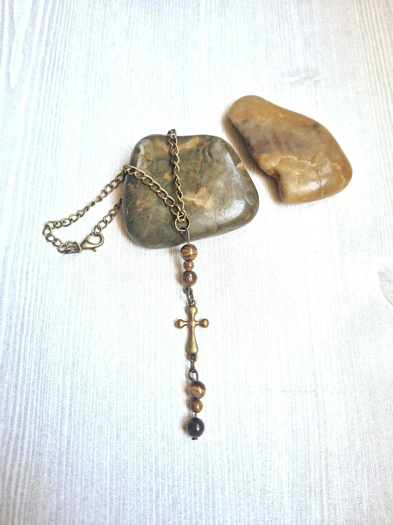May include: A necklace featuring a bronze-toned metal chain and a small cross pendant. The design includes several round, brown, and tan tiger eye beads arranged above and below the cross, finished with a lobster claw clasp closure.