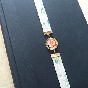May include: A white ribbon bookmark with a floral pattern and a round glass charm featuring a pink rose. The charm is attached to the ribbon with a gold metal connector.