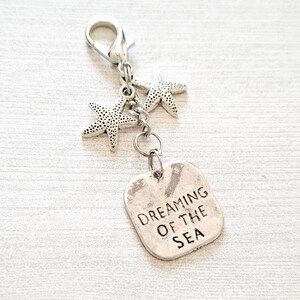 May include: A silver-toned charm with a lobster clasp, two starfish charms, and a rectangular tag. The tag is engraved with the words "DREAMING OF THE SEA". The charm is designed to be attached to a keychain or bag.