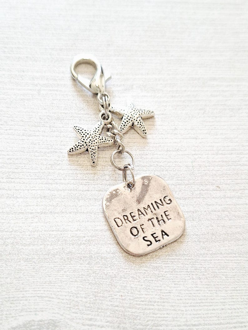 May include: A silver-toned charm with a lobster clasp, two starfish charms, and a rectangular tag. The tag is engraved with the words "DREAMING OF THE SEA". The charm is designed to be attached to a keychain or bag.