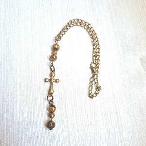 May include: A necklace featuring a bronze-toned metal chain with a lobster clasp, accented by a series of smooth, round, brown and gold tiger's eye beads and a central bronze-toned metal cross pendant hanging vertically from the chain.