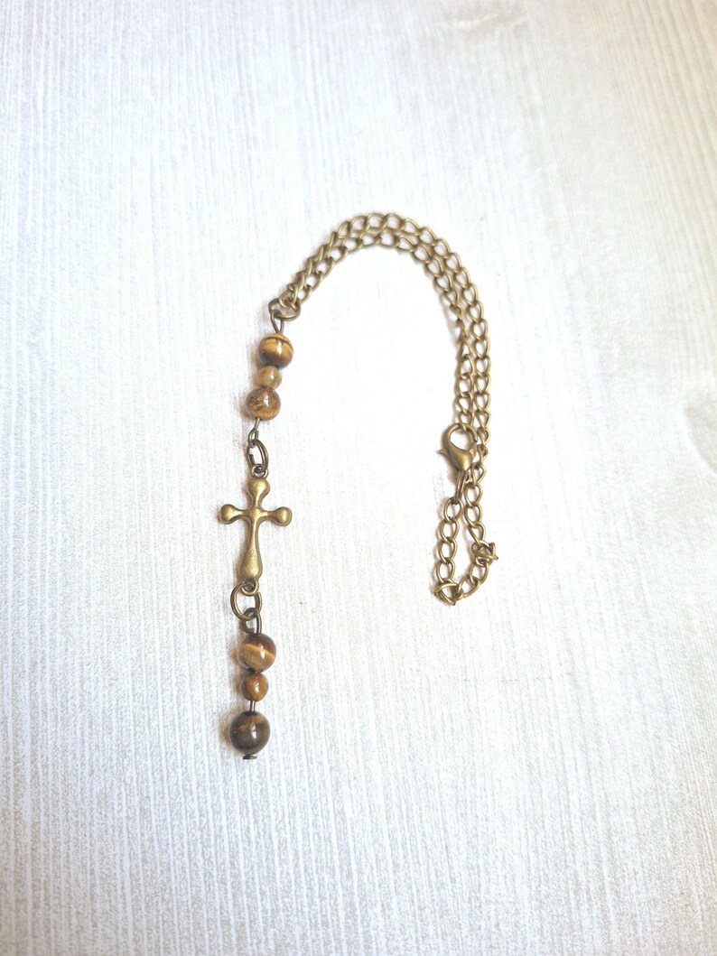 May include: A necklace featuring a bronze-toned metal chain with a lobster clasp, accented by a series of smooth, round, brown and gold tiger's eye beads and a central bronze-toned metal cross pendant hanging vertically from the chain.