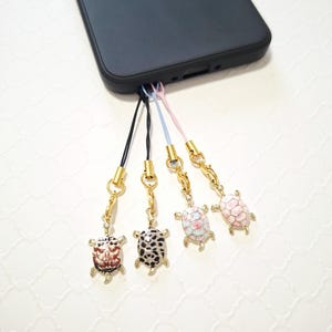 Pode incluir: Quatro berloques em forma de tartaruga presos a uma capa de smartphone preta por cordões finos. Os berloques apresentam ferragens de metal dourado e cascos de esmalte com vários padrões, incluindo vermelho e branco, preto e branco e rosa.