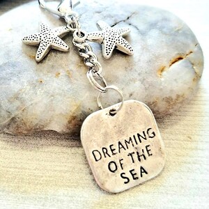 May include: A silver-toned keychain featuring two starfish charms and a rectangular tag. The tag is engraved with the words "DREAMING OF THE SEA" in black capital letters. The keychain is attached to a lobster clasp.