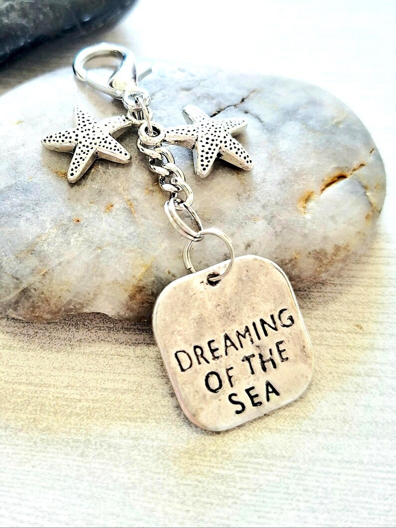 May include: A silver-toned keychain featuring two starfish charms and a rectangular tag. The tag is engraved with the words "DREAMING OF THE SEA" in black capital letters. The keychain is attached to a lobster clasp.