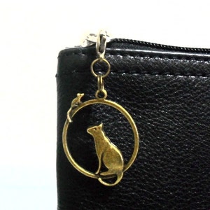 May include: A gold-toned charm featuring a cat and a mouse inside a circle. The charm is attached to a black leather zipper pull.