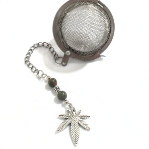 May include: A silver tea infuser with a chain and a green and brown stone bead. The chain is attached to a silver marijuana leaf charm.