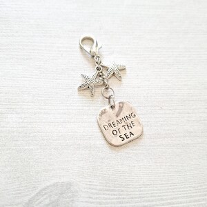 May include: Silver-toned charm featuring two starfish and a rectangular tag. The tag is engraved with the words "Dreaming of the Sea." The charm has a lobster clasp for easy attachment to a keychain or bag.