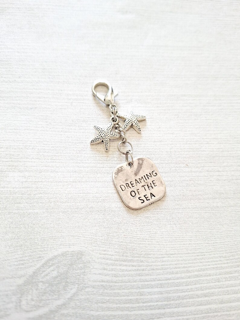 May include: Silver-toned charm featuring two starfish and a rectangular tag. The tag is engraved with the words "Dreaming of the Sea." The charm has a lobster clasp for easy attachment to a keychain or bag.