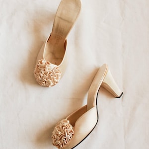 Op de afbeelding: Een paar vintage crèmekleurige muiltjes met hoge hakken. Elke schoen heeft een geplooid stoffen detail op de neus en een kleine, gestapelde hak. De schoenen staan tegen een neutrale achtergrond.