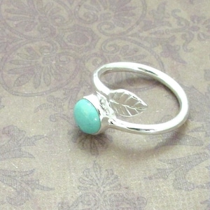 Puede incluir: Un anillo de plata con una piedra turquesa y un diseño de hoja. El anillo presenta una piedra redonda de color azul claro engastada en una banda de plata. Un pequeño detalle de hoja está incorporado en el diseño del anillo.