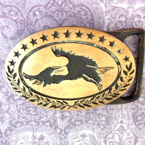 May include: Oval-shaped belt buckle with a brass finish. The buckle features a central design of a flying eagle in black, surrounded by stars and laurel leaves. The buckle has a dark bronze-colored frame.