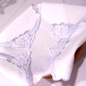 Op de afbeelding: Witte linnen servetten met een delicate druiven- en bladerborduursel in lichtblauw. De servetten hebben een geschulpte rand en zijn gevouwen, wat het gedetailleerde ontwerp laat zien. De servetten liggen in een houten mand.