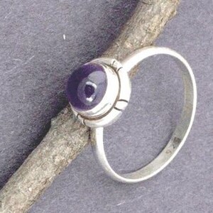 May include: A silver ring with a purple amethyst gemstone set in a bezel setting. The ring has a simple band and a delicate design.