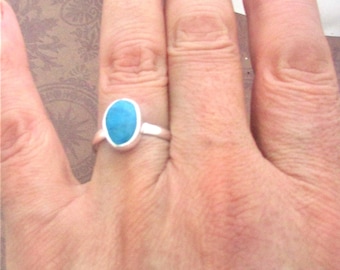 May include: A silver ring featuring a smooth, oval-shaped, bright turquoise stone set in a simple bezel. The band is plain and metallic, with a polished finish that complements the vibrant blue color of the central stone, creating a clean look.