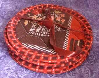 May include: A stack of three circular fabric coasters tied together with a thin brown ribbon. The coasters feature a brown background with a floral pattern and a red and white checkered border. The fabric is quilted with visible stitching.