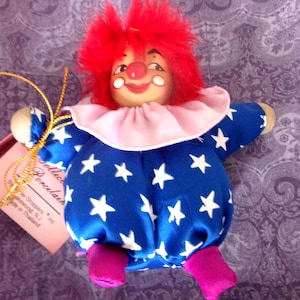 May include: A vintage clown doll with a red, fluffy wig and painted facial features. The doll wears a blue satin outfit with white stars, a pink ruffled collar, and magenta feet. A gold string and tag are attached.