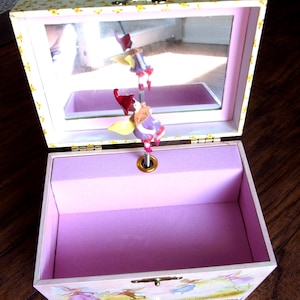 K&ouml;nnte beinhalten: Eine rosa Musik-Schmuckschatulle mit einem Spiegeldeckel und einer Feenfigur. Die Schatulle hat eine hellrosa Innenausstattung mit einem lavendelfarbenen Samtfutter. Das &Auml;u&szlig;ere zeigt eine Feenszene und einen goldfarbenen Verschluss.