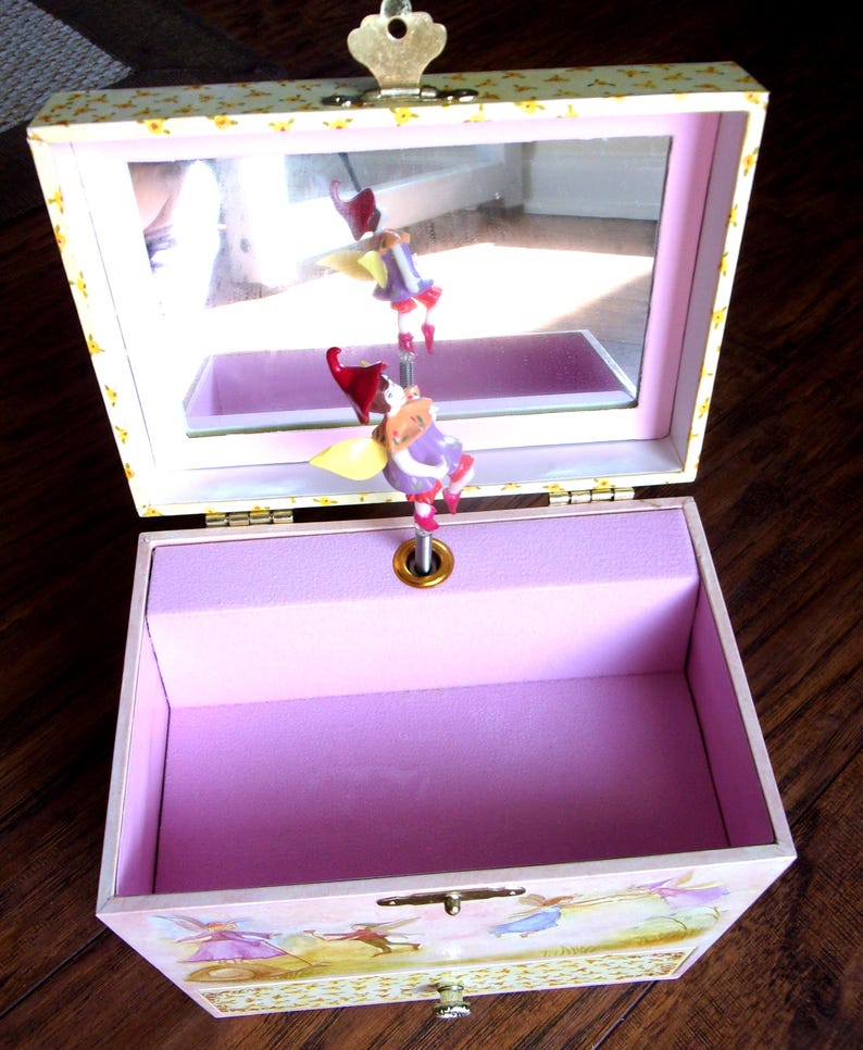 K&ouml;nnte beinhalten: Eine rosa Musik-Schmuckschatulle mit einem Spiegeldeckel und einer Feenfigur. Die Schatulle hat eine hellrosa Innenausstattung mit einem lavendelfarbenen Samtfutter. Das &Auml;u&szlig;ere zeigt eine Feenszene und einen goldfarbenen Verschluss.
