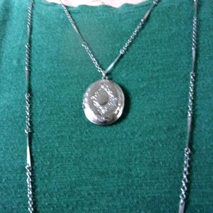 May include: Silver-toned locket necklace with a decorative design. The round locket hangs from a delicate chain, complemented by a longer chain with bar accents. The necklace is displayed against a green background.