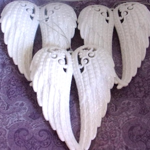 May include: Three pairs of white, glitter-covered angel wing ornaments. Each pair features detailed, moulded feather textures and an intricate, open-work scroll design at the top. Each wing ornament includes a thin silver hanging string.