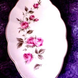 Peut inclure: Un plat de service ovale en porcelaine blanche avec un bord doré. Il est orné d'un motif floral de roses roses et de feuilles vertes. Les roses sont représentées à différents stades de floraison, ajoutant de la profondeur.
