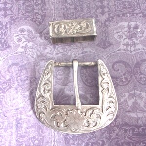 May include: Silver-toned metal belt buckle and matching keeper. The buckle is a horseshoe shape with floral and scrollwork designs. The keeper is rectangular with a floral design. The pieces are set against a patterned purple background.
