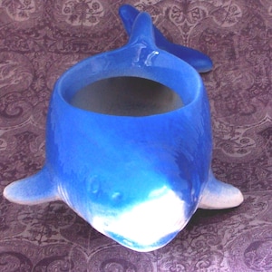May include: A ceramic whale-shaped planter in shades of blue, with a large opening for plants. The whale has a glossy finish with white detailing on the fins and belly. The planter is designed to stand upright.