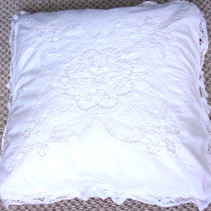 Puede incluir: Cojín cuadrado blanco con intrincado bordado floral en el centro. El cojín tiene un ribete de encaje alrededor de los bordes. Se encuentra sobre una superficie de color neutro.