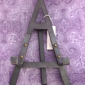 Puede incluir: Un caballete de metal gris oscuro con un diseño de tres patas. El caballete tiene dos tiras de metal grises con remaches de color latón. Una etiqueta blanca está sujeta a la parte superior del caballete con una cuerda blanca.