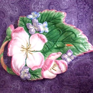May include: A ceramic decorative leaf-shaped dish with raised floral details. The dish features a large green leaf with pink and white flowers, and small purple and blue flowers. The dish has a glossy finish and is set against a purple background.