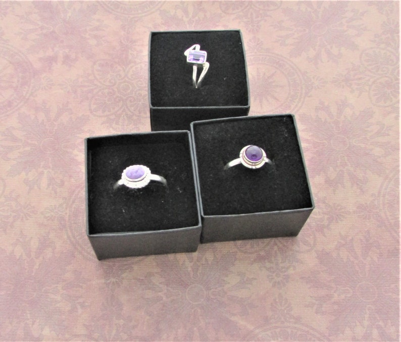 May include: Three small black ring boxes with purple amethyst rings inside. The rings are set in silver.