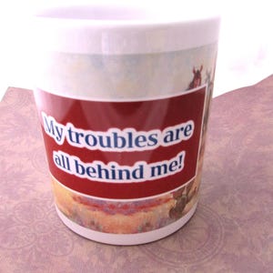 Op de afbeelding: Witte keramische mok met een rode band met de tekst "My troubles are all behind me!". De mok heeft een landschapsontwerp met paarden en een lichtblauwe en witte bovenkant.