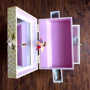 K&ouml;nnte beinhalten: Eine Vintage-Schmuckschatulle mit floralem Muster auf der Au&szlig;enseite. Die Box ist ge&ouml;ffnet und zeigt ein rosa Interieur, einen Spiegel und eine kleine Schublade. Eine dekorative Figur ist innen angebracht. Die Box ist aus Holz und hat einen goldfarbenen Verschluss.