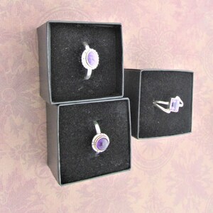 May include: Three small black boxes with velvet lining, each containing a silver ring with a purple gemstone.